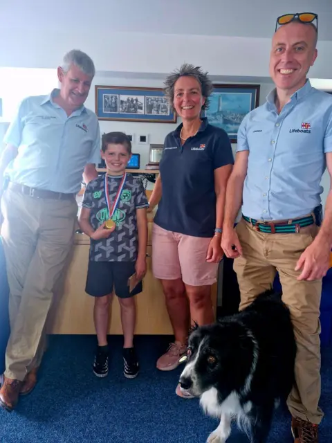 Barry Dock RNLI Marley smiles after being given his medal by three RNLI crew members, two men and a woman. A black and white sheepdog stands between the legs of a crew member.