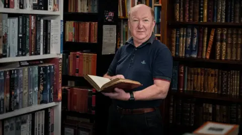 @kim.dungduong Christopher is stood in a darkened shop full of books. Some of them look old. He has short ginger hair and is looking at the camera. He has a book open in his hand.