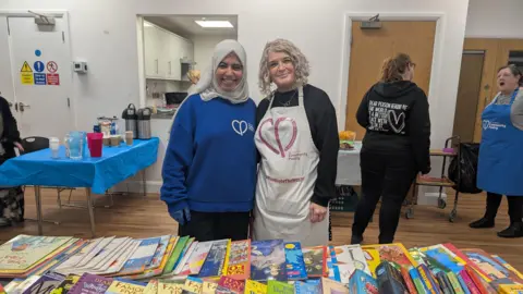 Tara Dolby/BBC Two smiling woman stood behind a table covered in children's books. One is wearing a black jumper and white apron. The other woman is wearing a blue jumper and white headscarf. 
