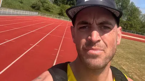 Joe Baker takes a selfie on a red athletics track. He is wearing a black baseball cap and a black running vest with a yellow running vest underneath. It is sunny and there is grass in the middle of the track.