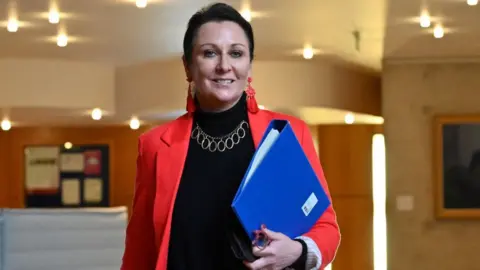 Getty Images Siobhian Brown, who has dark hair tied back, walks towards the camera. She is wearing a red jacket over a black turtleneck jumper, with a blue folder tucked under her arm. 