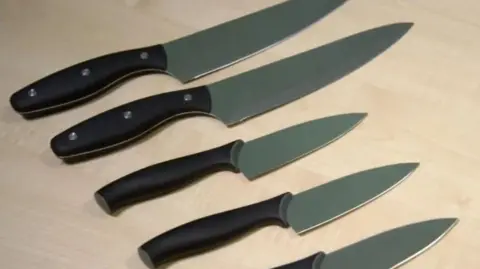 Five kitchen knives with black handles and silver blades lie on a wooden worktop.