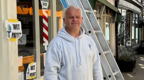 Matthew has white hair and is wearing a grey hooded top. He's standing by a shop which he's painting. 