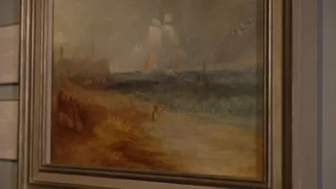 A Turner oil landscape painting of a ship just offshore. The beach and dunes are orange, russet, yellow and cream, while the sea is many shades of blue. The ship's sails are pure white against a grey-white sky.