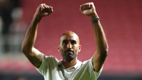 Walsall head coach Lee Grant raises both arms in celebration to the club's fans after their EFL Cup win over Bristol City. He is wearing a cream coloured t-shirt.