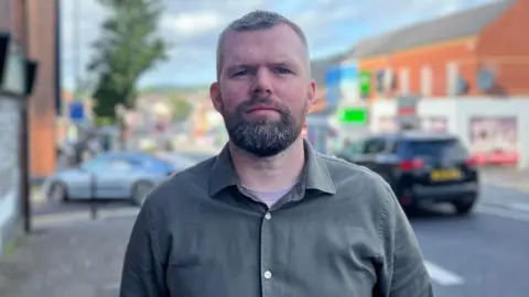 Gerry Carroll standing on street with traffic in the background. He is wearing a dark green shirt over a purple t-shirt. He has short dark and greying hair with a beard.