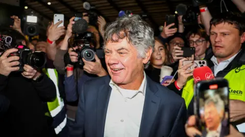 Reuters Gerry Hutch, who is an older man with dark, greying hair is surrounded by a media scrum as he enters a count centre in Dublin. He is wearing a dark jacket and white collared shirt.