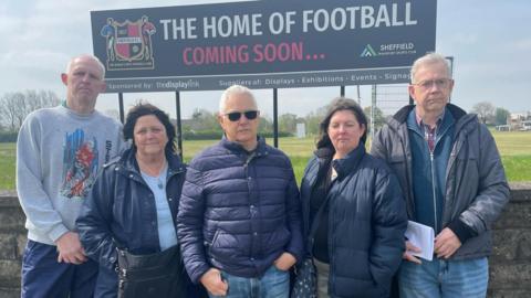 Three men and two women look cross as they stand infront of a large sign advertising the 'home of football'