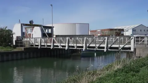 A bascule bridge over murky water. Industrial buildings can be seen in the distance. The sky is blue.