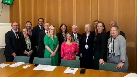 Sarah Hall Members of the APPG on autism standing in a board room against a wooden wall and green chairs. There is a table in front of them with various papers laid out on it.