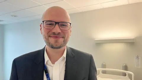 ELLEN KNIGHT/BBC Ned pictured smiling and looking into the camera. He's wearing a white collared shirt and a dark grey suit jacket. He's stood in one of the hospital rooms, which has white walls and a white ceiling. 