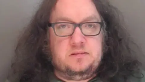 Durham Police Mugshot of Madgin. He has a fat face with a scruffy grey goatee beard and thick shoulder length frizzy dark hair. He is also wearing glasses.