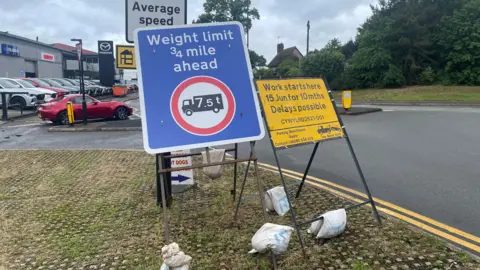A yellow roadworks sign reads "Work starts here 15 Jun for 10 mths. Delays possible." It stands next to a blue weight limit sign on a grass verge beside a road. A car dealership and trees are visible in the background.