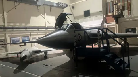 Photograph of a prototype Vulcan bomber - alongside a model of a Vulcan bomber. The image shows the aeroplanes in a storage warehouse.