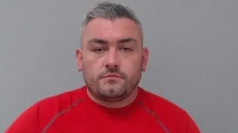 PSNI Brendan O'Callaghan mug shot. He has grey short hair and dark stubble. He is wearing a red t-shirt. 