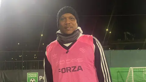 Karim Suliman wrapped up warm for playing football in the cold and dark. He had a hat and scarf on and is wearing a pink vest. There is a goal behind him. 