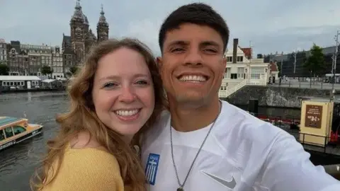 A man wearing a white football top smiling with his arm around a woman wearing a yellow top. Behind them is a city scene.