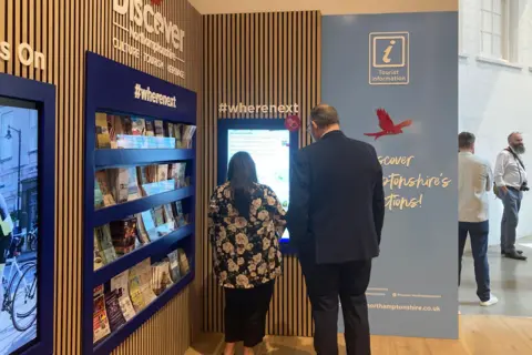 BBC The back of a tall man in a dark suit and a person with long hair and a floral top, as they look at the new digital screen at the information point indoors.