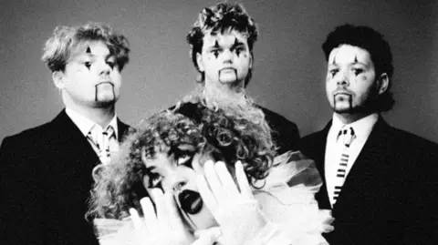 Supplied A black and white press image of She's In Parties. The lead singer is at the front of the image while her three fellow band members are standing behind her. They all have a clown face paint on.
