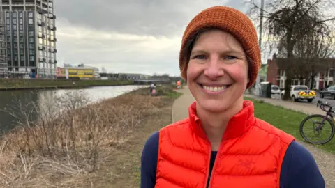 A woman wearing an orange beanie hat, orange gilet and a navy top. She's standing on a footpath along the river Taff.