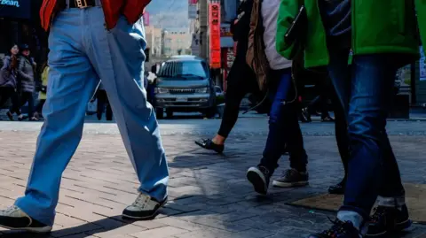A close-up shot of the lower bodies of men wearing denim jeans walking on a busy street in South Korea. 