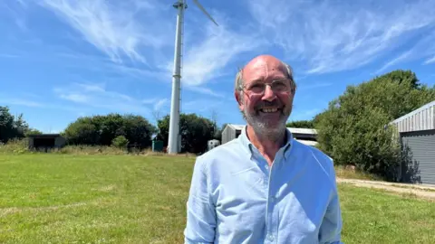 Pete West in a blue shirt in a field with a wind turbine in the background.