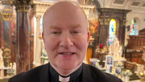 A priest in a church. He wears a black jacket and white dog collar.
