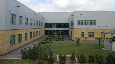HM Inspectorate of Prisons The outside of a prison, with planting. There is one main block and a grassed area. 