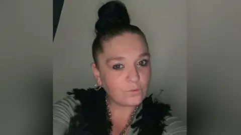 A woman with dark hair in a bun wears grey and black and looks at the camera taking a selfie.