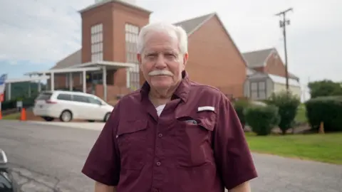 Eloise Alanna Seventy-five-year-old Willam Bogle, sporting white hear and a full white mustache, wears a maroon button-up shirt as he stands in front of a brick church 