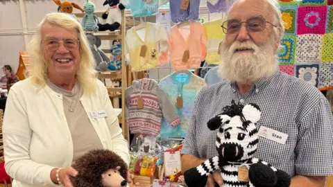 Hagley Village Market An older man and women smile as they hold up a knitted hedgehog and zebra in front of a stand at a craft fair. Behind them are knitted baby cardigans and a crocheted blanket hanging on a wooden frame. the woman has shoulder length blonde hair and is wearing silver wire-famed glasses, a cream cardigan and beige t-shirt. The man has a white beard and hair, is wearing tinted glasses, and a beige checked shirt.