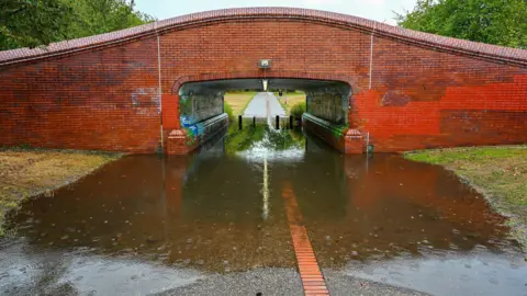 SMUpix/BBC Weather Watchers A red bricked bridge goes across a pathway. Large volumes of water has caused the underpass to flood and has started to travel across grass areas either side of the path.