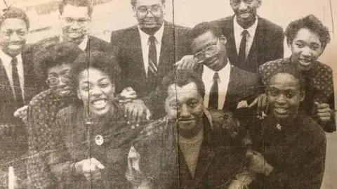 Birmingham Evening Mail Um recorte de jornal mostra o cantor com os membros do coro em uma foto em preto e branco. O cantor está parado no meio com os coristas em pé ao seu redor, segurando seus braços e sorrindo.