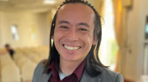 A close up of Deputy David Goy smiling. He has long dark hair and is wearing a red shirt with a grey jacket.