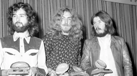 Press Association Three men in a black and white image hold awards against a striped background. On the left, the man has curly dark hair and a beard with a cowboy style shirt. The man in the middle has long blond wavy hair and a beard and wears a dark shirt with white flowers on it. The man on the right has straight shoulder length brown hair and a brown beard and is wearing a white shirt and leather jacket.