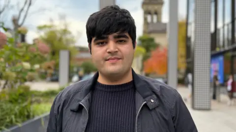 Queens University student Jay Basra standing in front of the university's students' union - he is wearing a dark jacket and navy jumper. 
