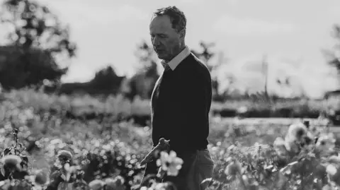 David Austin Roses A black and white photo of David Austin in the middle of a field of flowers, looking down at them. He is wearing dark trousers with a black jumper and a white collar