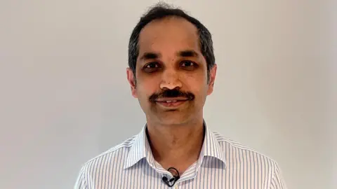 A man is standing in front of a white wall. He has tan skin and black hair. He also has a black moustache. He is wearing a white shirt with blue stripes. 