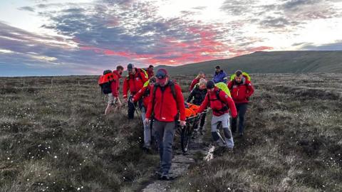 Several people in red jackets wheeling down a man on a stretcher down a mountain as the sun rises