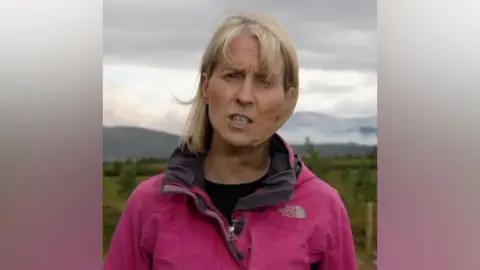Katie Hunter stands outside wearing a pink windproof coat, gesturing as she speaks to camera in front of heathland and cloudy sky in near Aviemore, Scotland on Saturday.