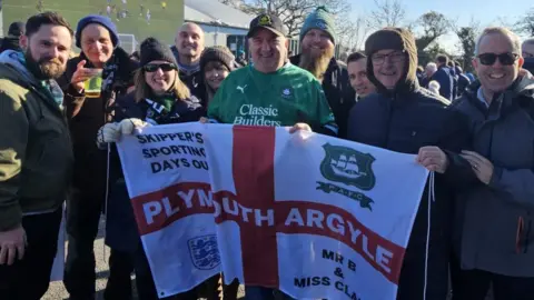 A group of Plymouth Argyle supporters from the North Devon Greens group stand outside a football stadium holding an England flag with the team's name, the England national team badge, Argyle badge and messages on it. A big screen showing another football match is in the background.