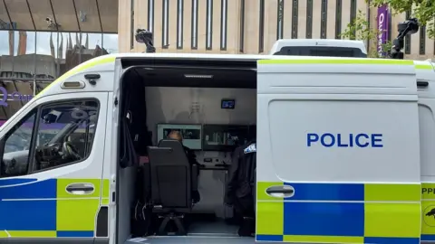 A white police van with green and blue squares on it and police written on it in blue. A police officer can be seen sitting inside the van - looking at a screen.