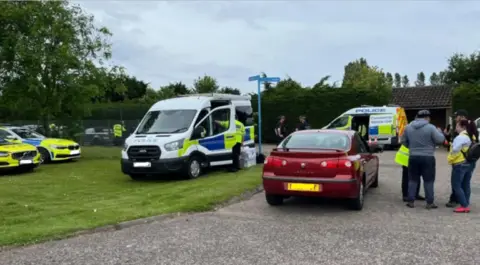 Andrew Turner/BBC A couple speak to police by their car with lots of police vehicles parked nearby