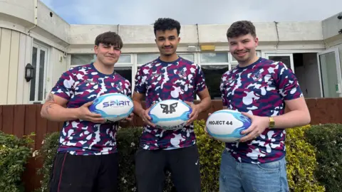 Handout Three young men are holding rugby balls while standing, looking at the camera in front of an old building with large windows.