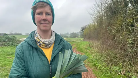 Ella Gent stand on a rural path in a field. She person is wearing a quilted green jacket, a yellow shirt underneath, a patterned neck scarf and a teal knitted hat. She is holding a bundle of long green leaves that appear to be freshly harvested leeks.