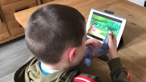 A young child with asthma holds his inhaler. The clear, colourless plastic device is electronically linked to a video game on a tablet in front of him.