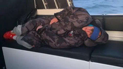 Oscar is asleep on a seat on a boat. He has a black camo puffer jacket on, a blue hat, grey trousers, white socks and red crocs. The sea is in the background.