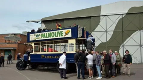 London Bus Museum A heritage open top bus painted in royal blue with vintage advertising hoardings is parked outside a green hangar with spectators queuing to board it