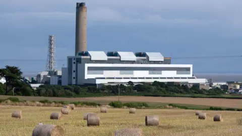 A wide-shot of Aberthaw Power Station, with a farmers' field and bales of hay in the foreground. Aberthaw is a white building with a chimney on its left hand side. A structure similar to a pylon is in the background and there is a blue sky.