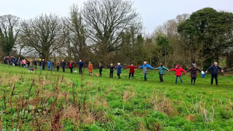 People for Packsaddle A line of people wearing coats hold hands across a field with trees in the background.